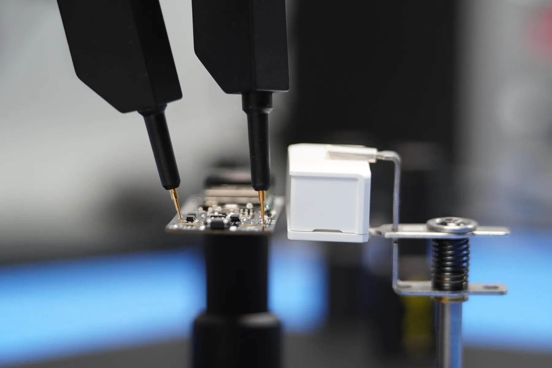 Two black test probes with gold tips touching the pads of a small OBJEX board held in a spring-loaded bench fixture